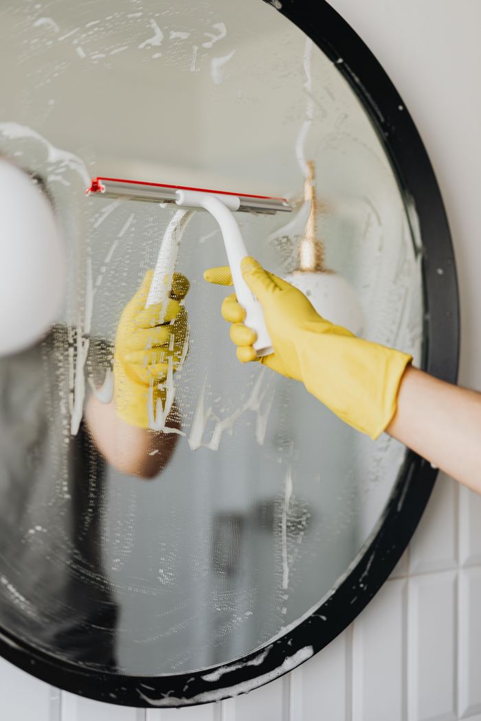 about-02 Faceless housekeeper in yellow rubber gloves cleaning round mirror from foam by squeegee in bathroom